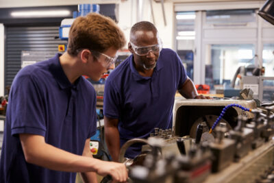 Two men wearing safety goggles and blue polo shirts work together on a machine in a workshop or factory setting.