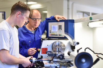 Two men wearing safety glasses operate a CNC milling machine in a workshop, with one providing instruction to the other.