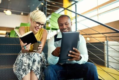 Two people sit on stairs, one holding a notebook and coffee, the other holding a tablet. They are smiling and appear to be engaged in conversation.