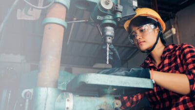 Woman wearing a hard hat and safety goggles operates a large industrial drill press in a workshop.