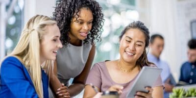 Three women are gathered around a tablet, smiling and engaged, in an office setting with others in the background.