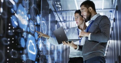 Two people look at a digital display with cloud and technology icons in a server room, holding a laptop and discussing data.