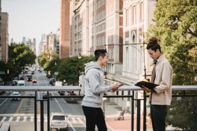 Two people stand on a city balcony. One holds an open folder while the other looks at it. A street with buildings and trees is in the background.