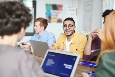 A group of people sitting around a table with laptops, engaging in discussion. One person in a yellow jacket is smiling and gesturing.