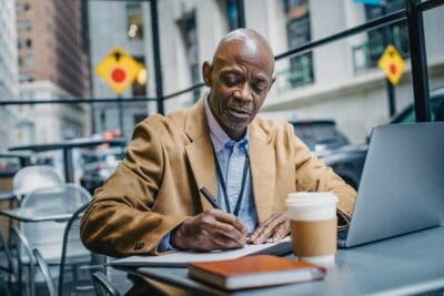 Man in a brown coat writes in a notebook at an outdoor cafe table with a laptop and coffee cup nearby.