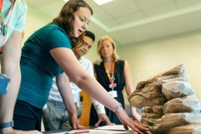A group of people examining a rock formation model on a table, with one woman pointing and others observing attentively.