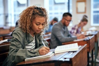 A woman sits at a desk in a classroom, writing in a notebook with an open textbook in front of her. Other students are seated in the background, also engaged in writing.
