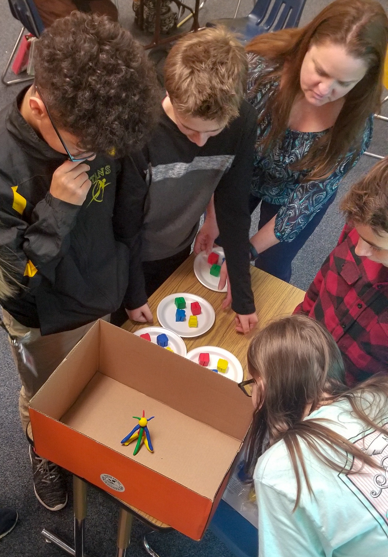 Students and a teacher gather around a table with a cardboard box and colorful clay figures on plates, appearing to work on a project together.