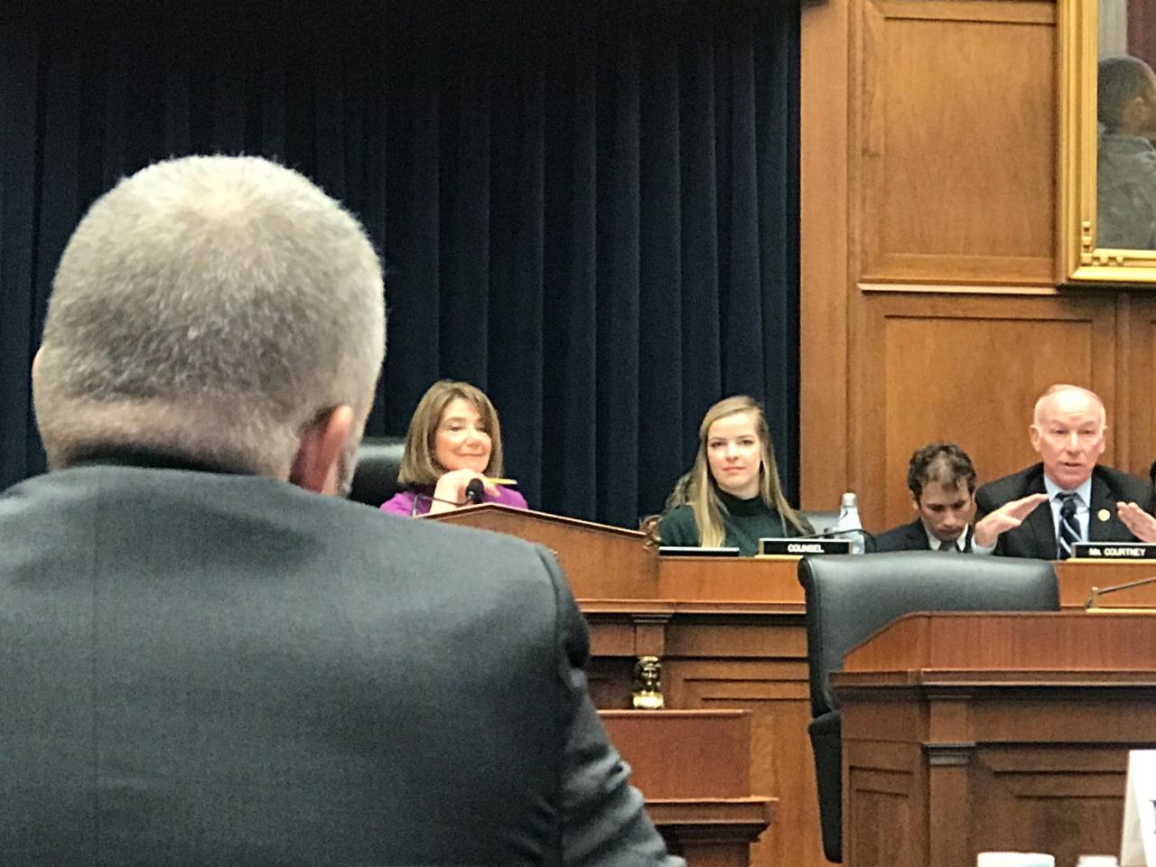 A panel of four people seated at a hearing table, with a speaker addressing them from the foreground.