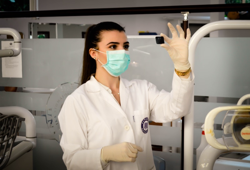 A woman in a lab coat and mask examines a small object in a laboratory setting.