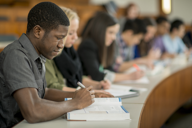 Students seated in a classroom are writing notes during a lecture.