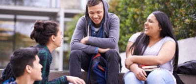 Four young adults sit outside, smiling and talking. One holds books, and another has a backpack. Trees and a building are in the background.