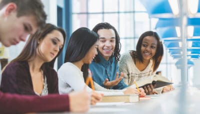 A group of students sits at a table, collaborating and taking notes, with books and papers spread out.