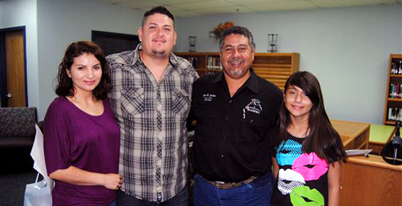Four people standing together indoors, smiling at the camera. A man and a woman are on the left, another man is in the center, and a young girl is on the right.