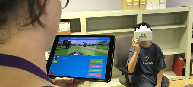A person holds a tablet displaying a game, while another person sits wearing a VR headset in a room with shelves.