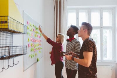 Three people in a room collaborate at a whiteboard covered with sticky notes, discussing and adding new ones.