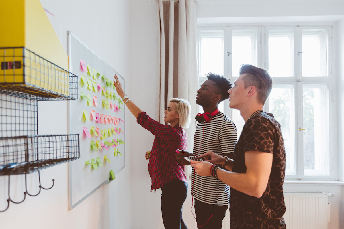 Three people in a room collaborate at a whiteboard covered with sticky notes, discussing and adding new ones.