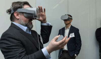 Two people wearing VR headsets stand in a room. One holds a controller while gesturing with his hand.