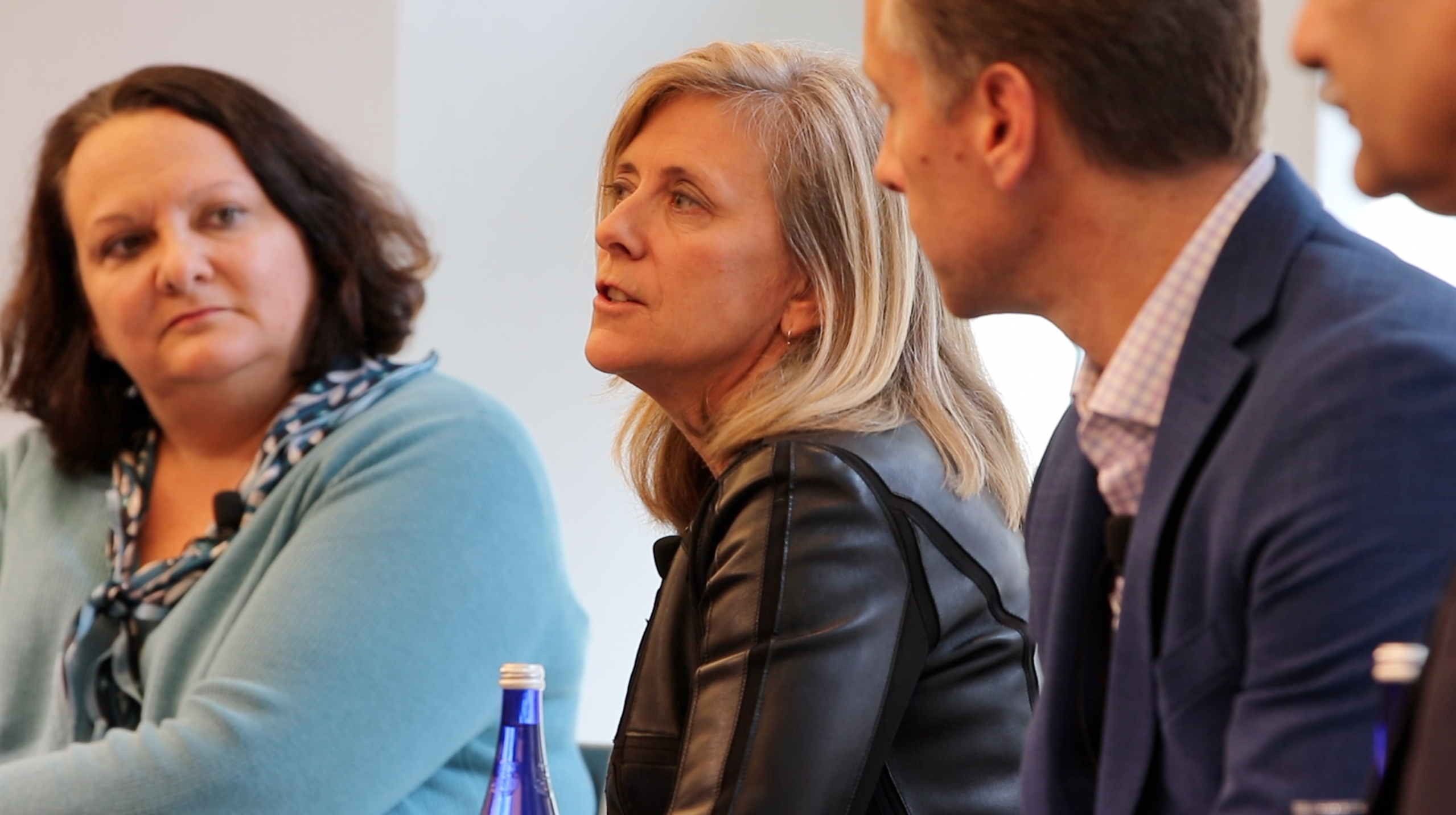 Three people engaged in a discussion, seated at a table. Two women and one man are visible, with focus on the woman in a black jacket speaking. Bottled water is on the table.