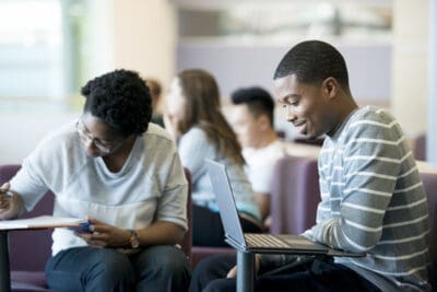 Two people sit on chairs in a room. One writes on paper, while the other uses a laptop. More people sit in the background.