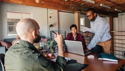 Three people discuss at a table with a laptop and notebooks in a modern office setting.