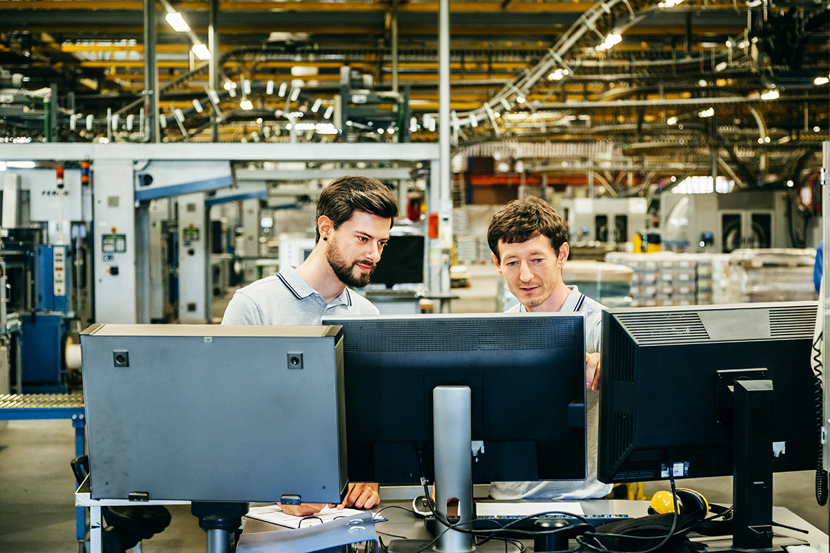 Two men are working at desks with computers in a factory setting, examining screens amid industrial equipment.