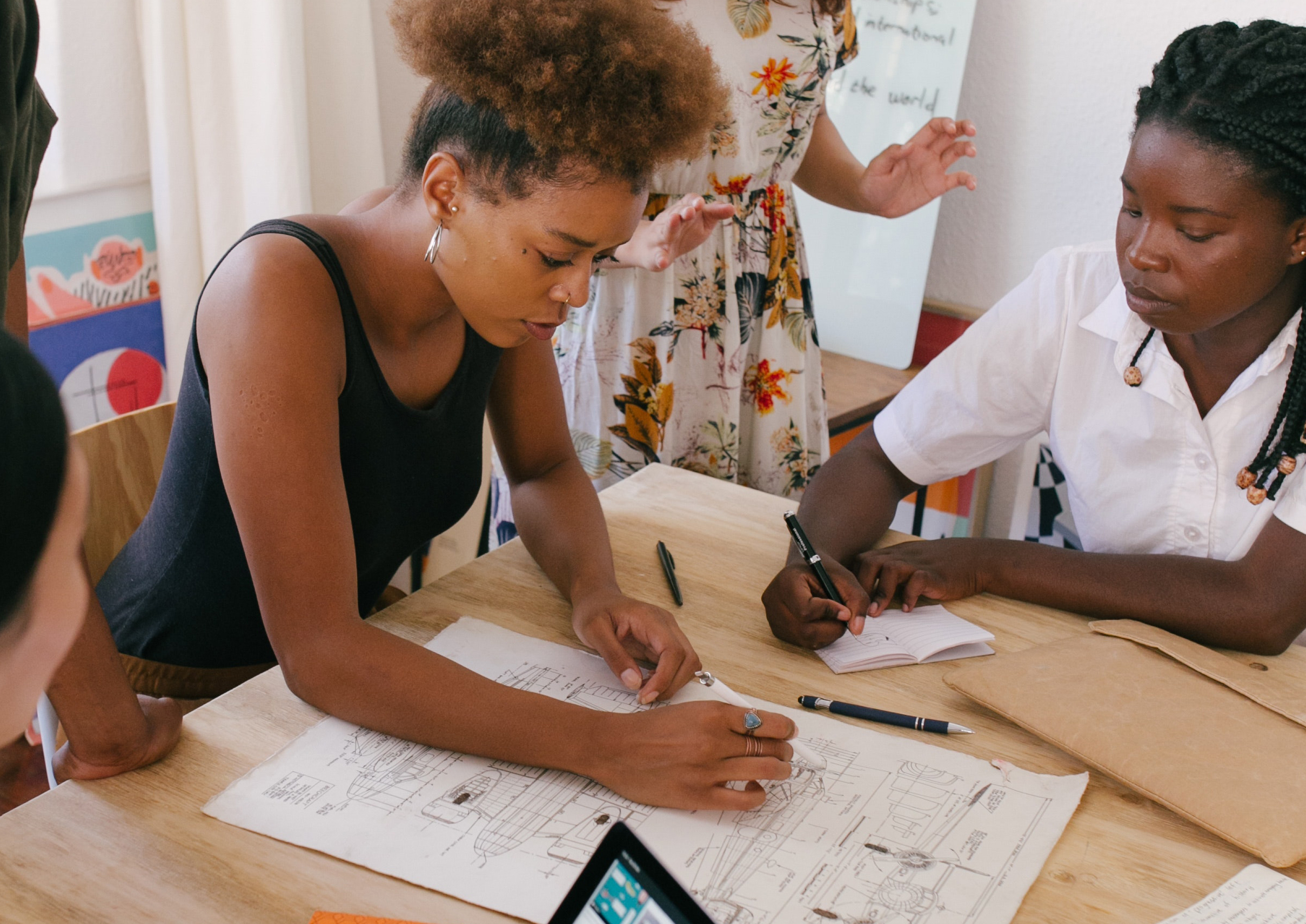 Two people sit at a table reviewing architectural drawings, surrounded by notebooks and a tablet.