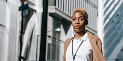 Person with short hair, wearing a white shirt and beige jacket, walks outdoors in an urban setting.