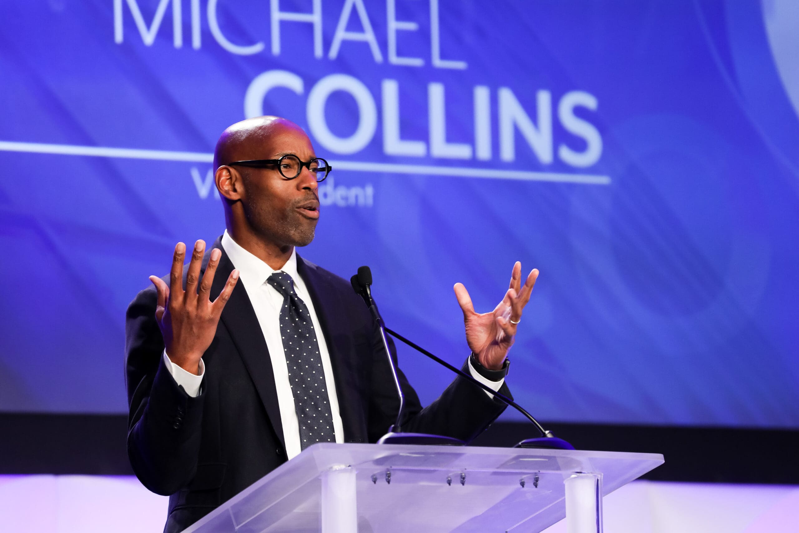 A man in a suit and tie speaks at a podium with a microphone against a purple backdrop displaying text.
