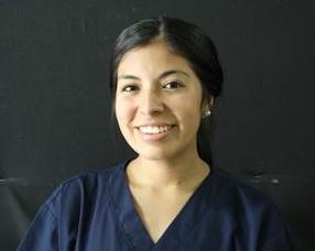 A person with long dark hair smiles, wearing a dark blue scrub top against a plain dark background.