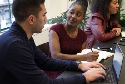 Three people are seated at a table. One person types on a laptop, another writes in a notebook, and the third listens. There are plants in the background.
