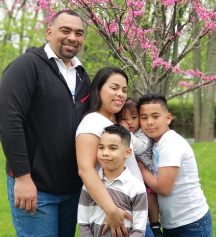 A family of five stands together outside near a blooming tree. The parents are with their three children, two boys and a girl, smiling at the camera.