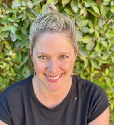 Person smiling in front of green foliage, wearing a black shirt with hair tied up.