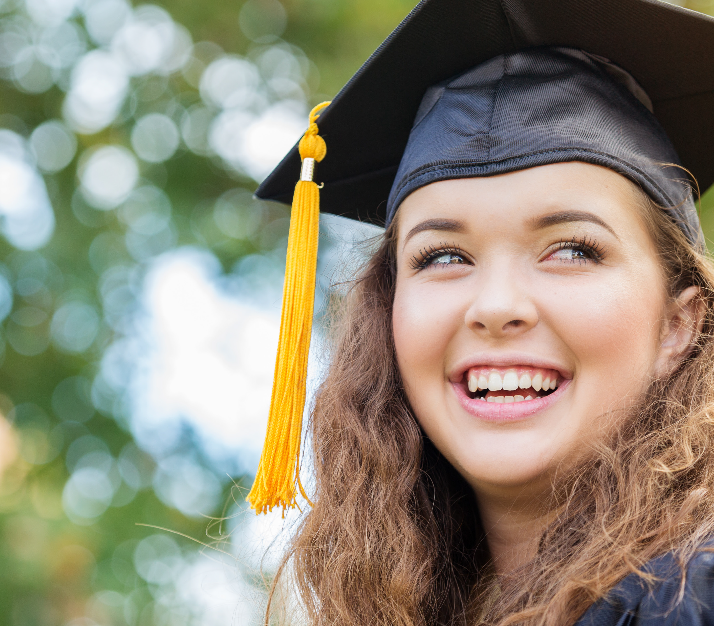 A smiling graduate wearing a cap and gown with a yellow tassel, standing outdoors with a blurred green background.