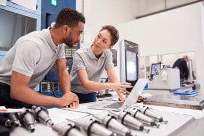 Two engineers in a workshop discuss plans on a laptop. Precision parts are laid out in the foreground on a table.