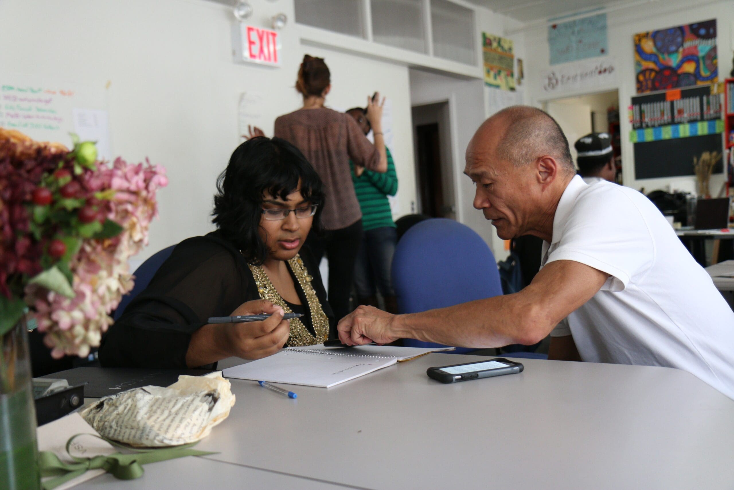 A man and woman collaborate at a table in an office, surrounded by papers and a cell phone. Other people are seen in the background.