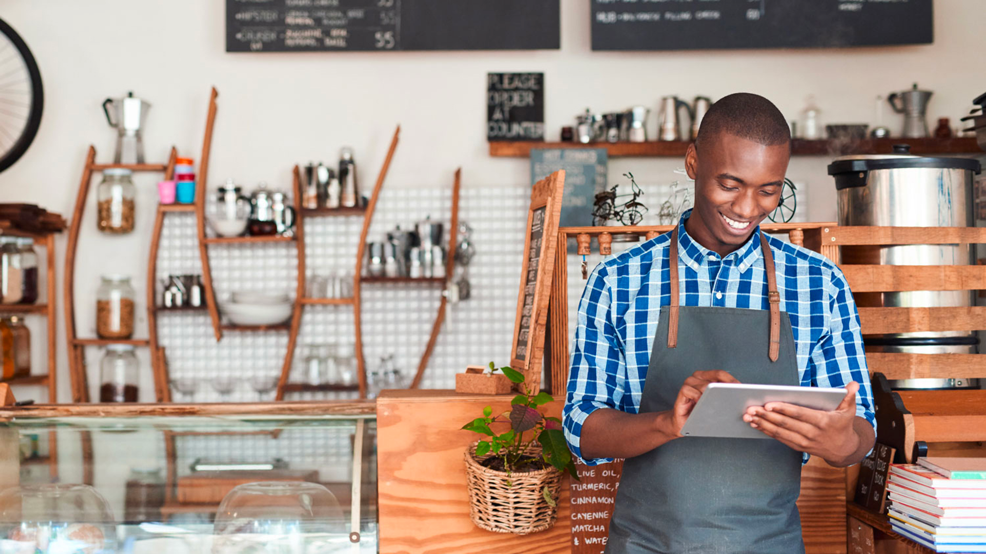 A barista in a blue checkered shirt and apron smiles while using a tablet in a modern coffee shop.