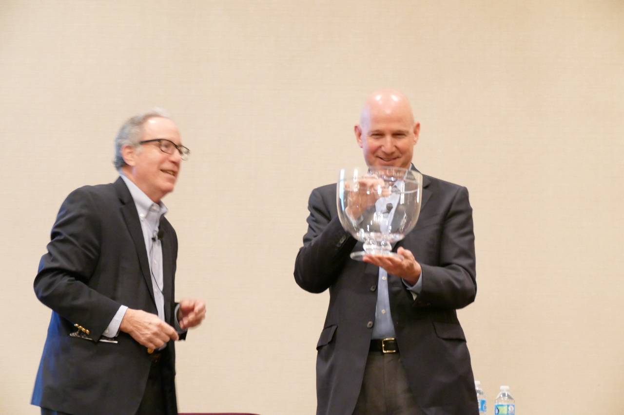 Two men in suits on stage; one holds a large clear glass award.