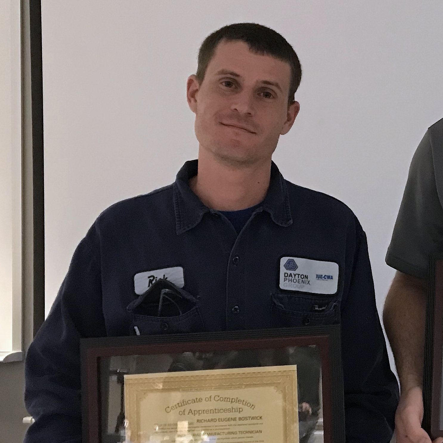 A person in a work uniform is holding a framed certificate of completion of apprenticeship.