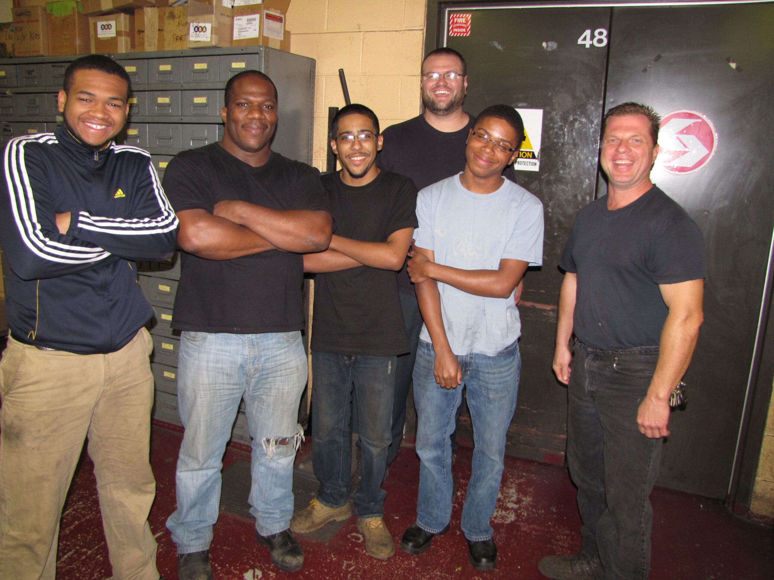 Six men standing together in a workplace environment, posing for a group photo. Some are smiling, wearing casual or work attire, with industrial cabinets and a door in the background.