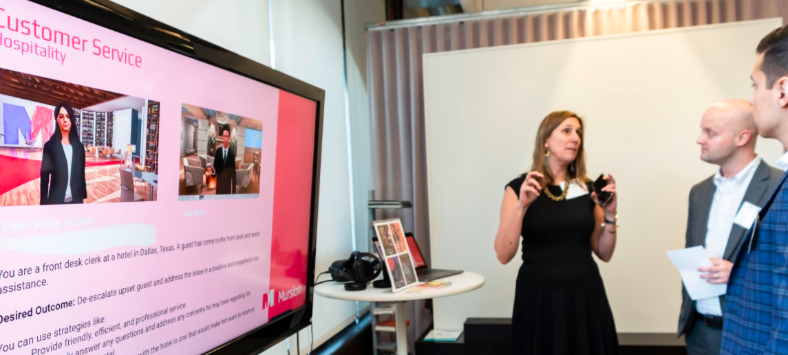A woman presents at a display screen during a business event, with two people listening and a table with a headset nearby.