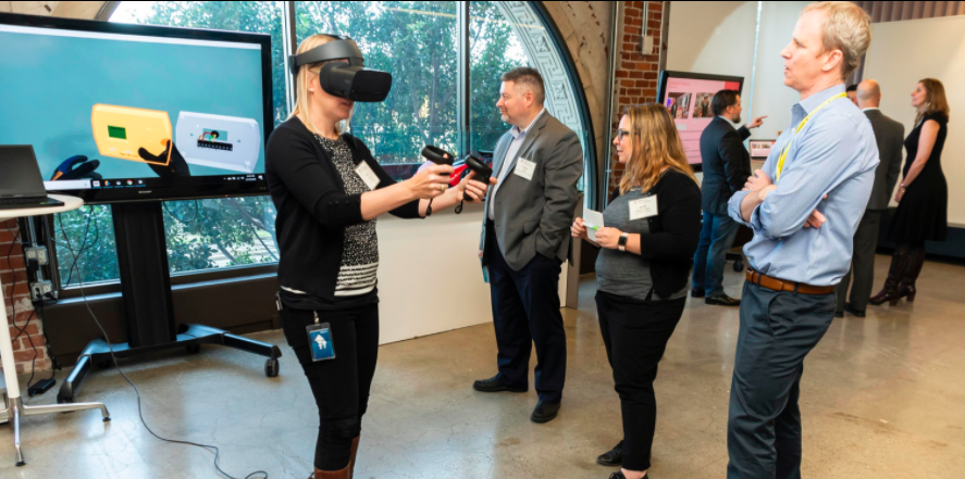 A person uses a virtual reality headset and controllers in a tech setup while others observe in an office setting.