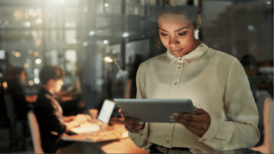 A woman in an office looks at a tablet while a person in the background works on a laptop.