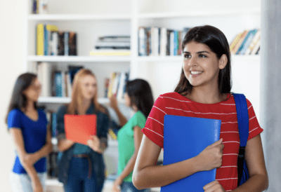 A woman in a red and white striped shirt holds a blue folder, standing apart from three others talking in the background. She smiles, standing by a bookshelf.