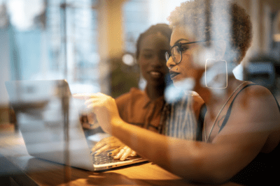 Two people are working together on a laptop in a sunlit room, with reflections visible on the glass surface.