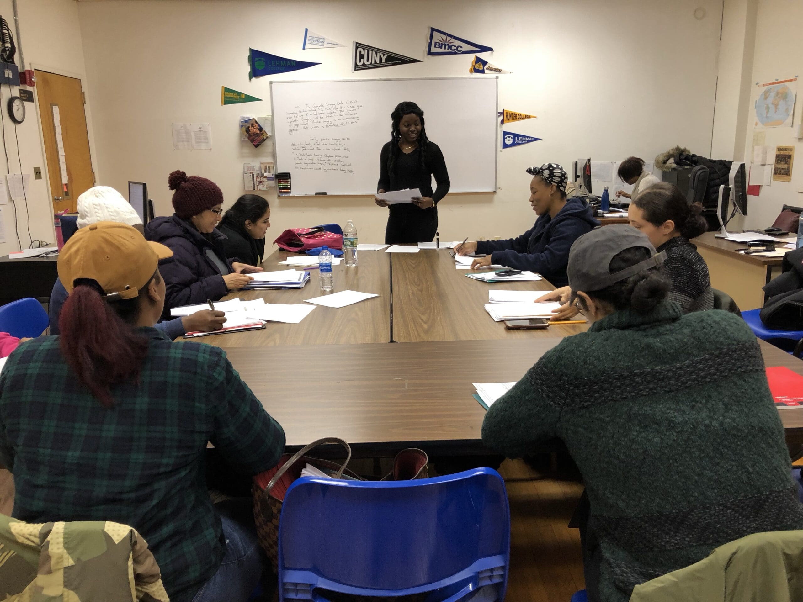 A group of adults sit around a table in a classroom, listening to a woman standing and speaking in front of a whiteboard.