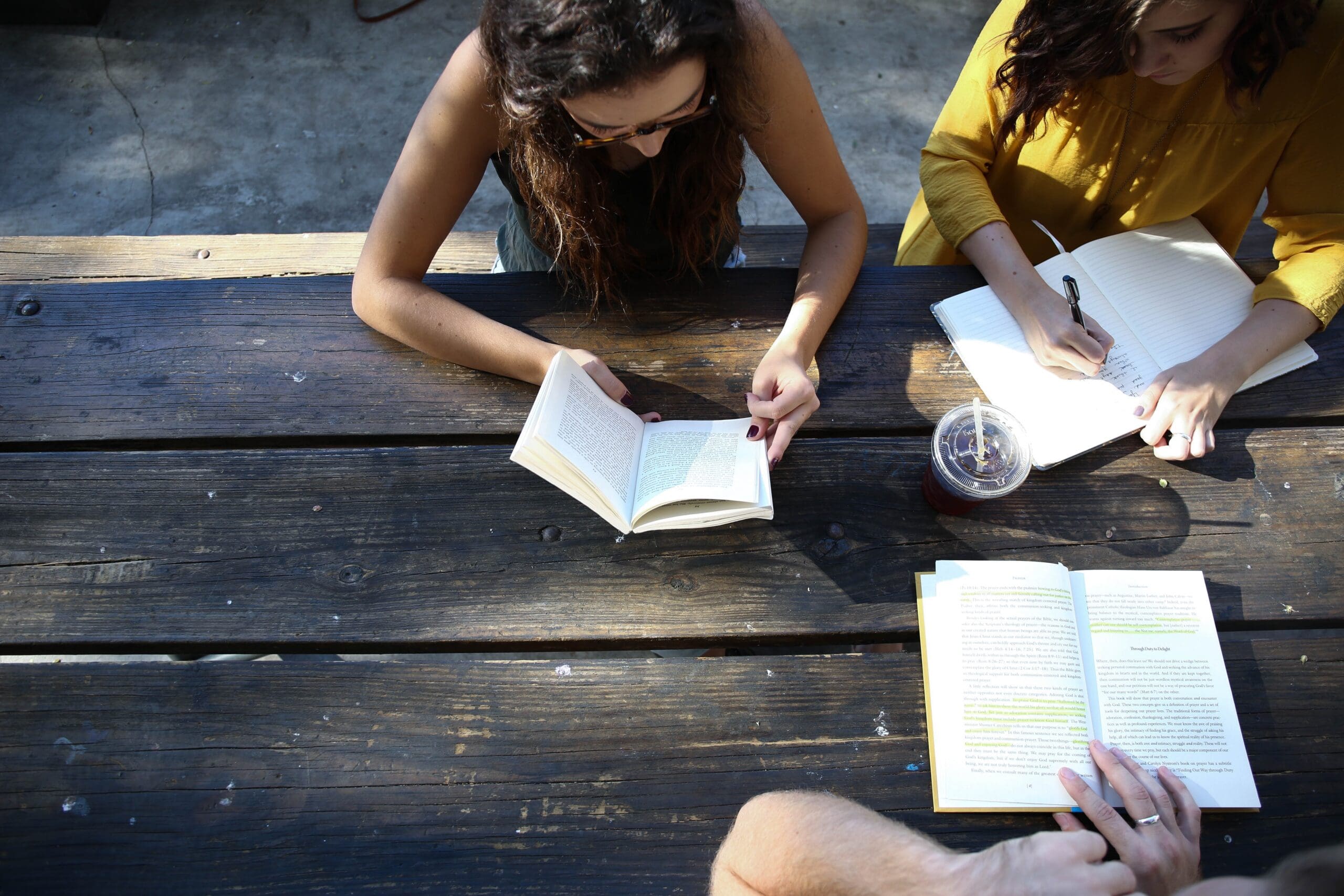 Three people sitting at a wooden table outdoors, reading books and writing notes. A plastic cup with a straw is on the table.