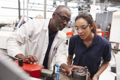 Two people in a workshop examine machinery. The man wears a lab coat, and the woman wears safety glasses and a navy shirt.