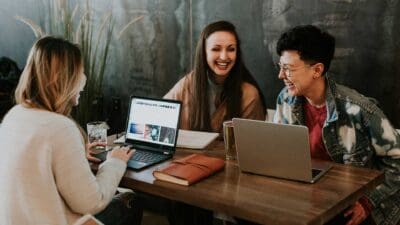 Three people sitting at a table with laptops, talking and smiling.