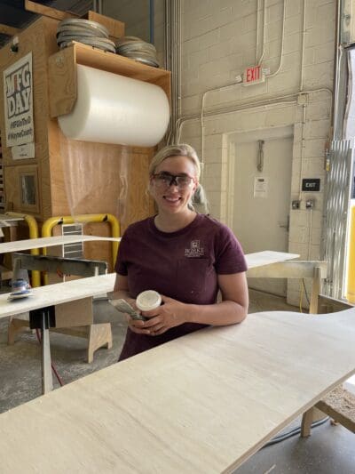 Person in a workshop holding a container, standing by a wooden table. Safety goggles are on their head. Various tools and materials are visible in the background.
